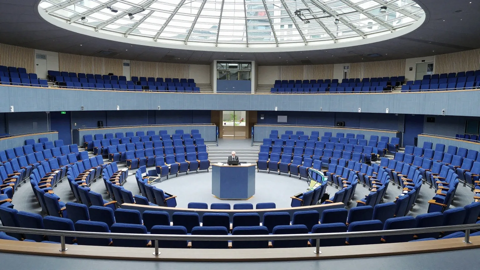 Ein großer Plenarsaal von der Tribüne aus, blaue Sitzreihen im Halbrund unter einer hellen Glasdecke